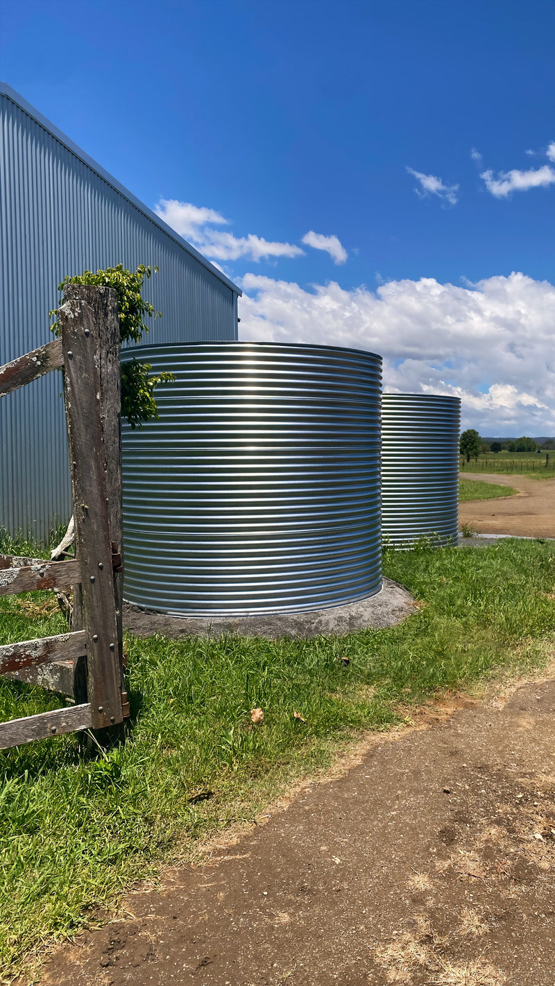 two large ccwt round water tanks on a concrete pad next to a big shed with an old fence in the foreground